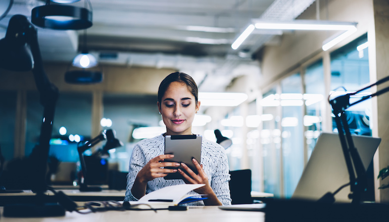 A woman sits at a desk holding a tablet while smiling at it.